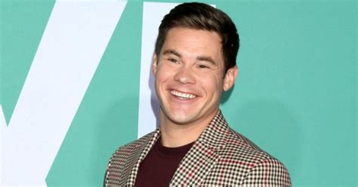 Adam Devine smiling in front of a gray background