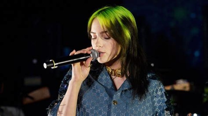 Billie Eilish singing into a microphone and holding one hand up with a finger raised