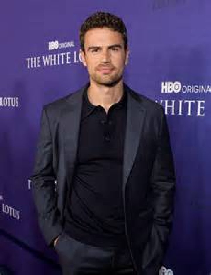 Theo James stands in front of greenery wearing a black suit